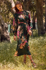 woman wearing a green floral midi dress with long sleeves and a belt, standing in a grassy area outdoors