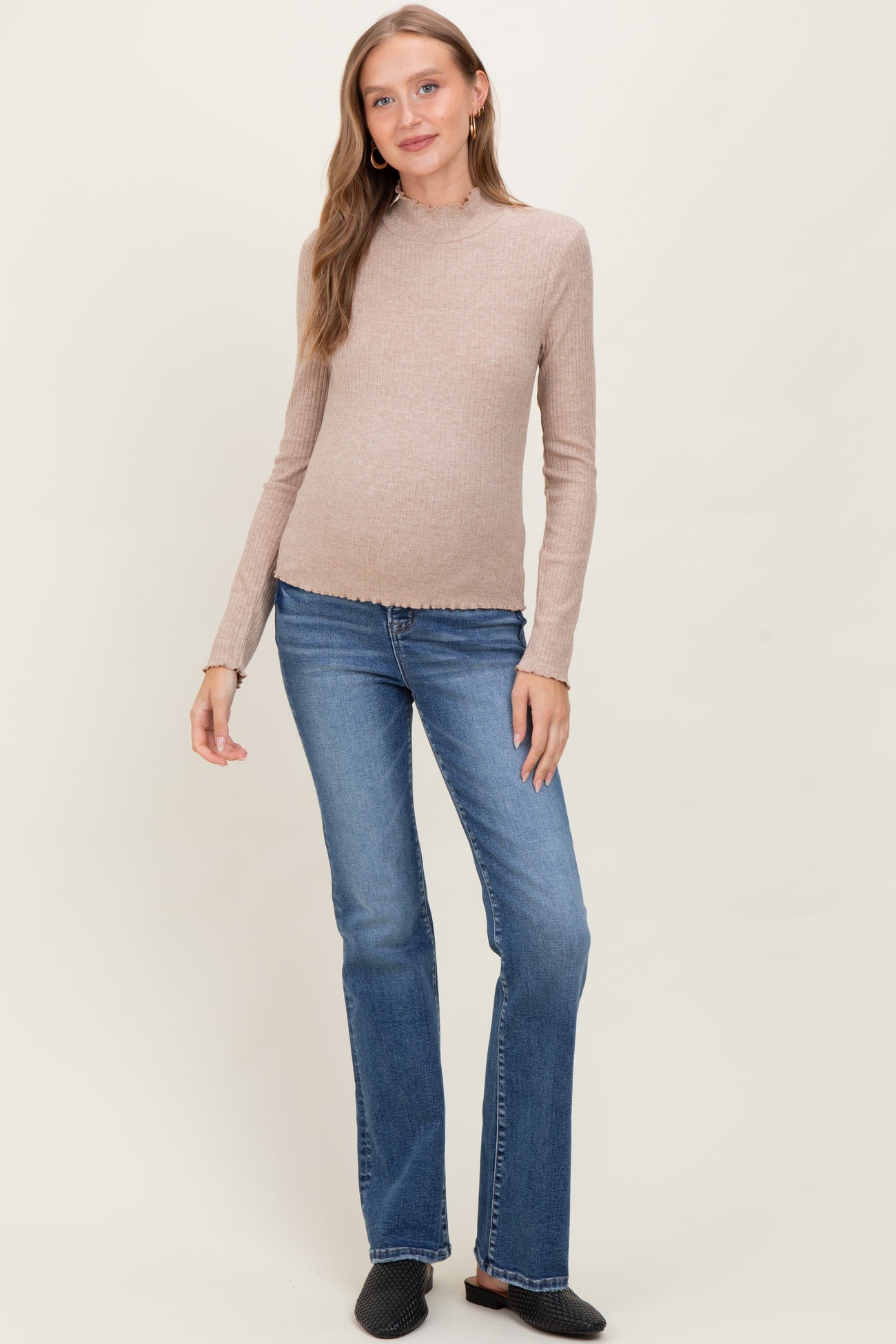 woman wearing a beige long sleeve maternity top with a scalloped hem and blue flared maternity jeans, showcasing a baby bump