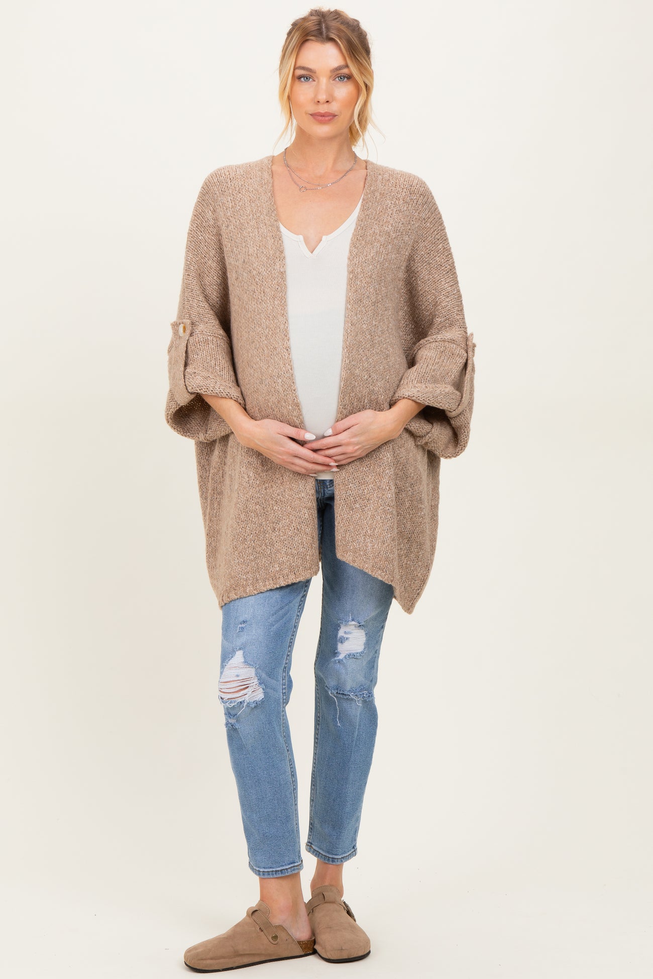 woman wearing a beige knitted maternity cardigan over a white top, holding her baby bump, paired with distressed jeans