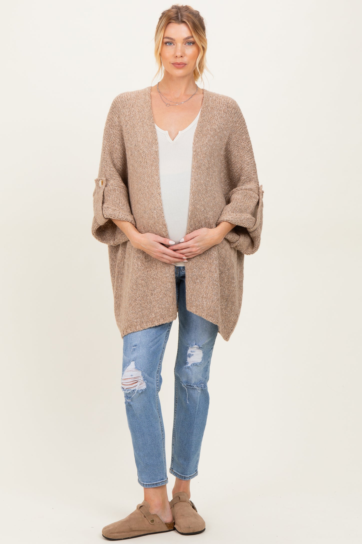 woman wearing a beige knitted maternity cardigan over a white top, holding her baby bump, paired with distressed jeans