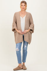 woman wearing a beige knitted maternity cardigan over a white top, holding her baby bump, paired with distressed jeans