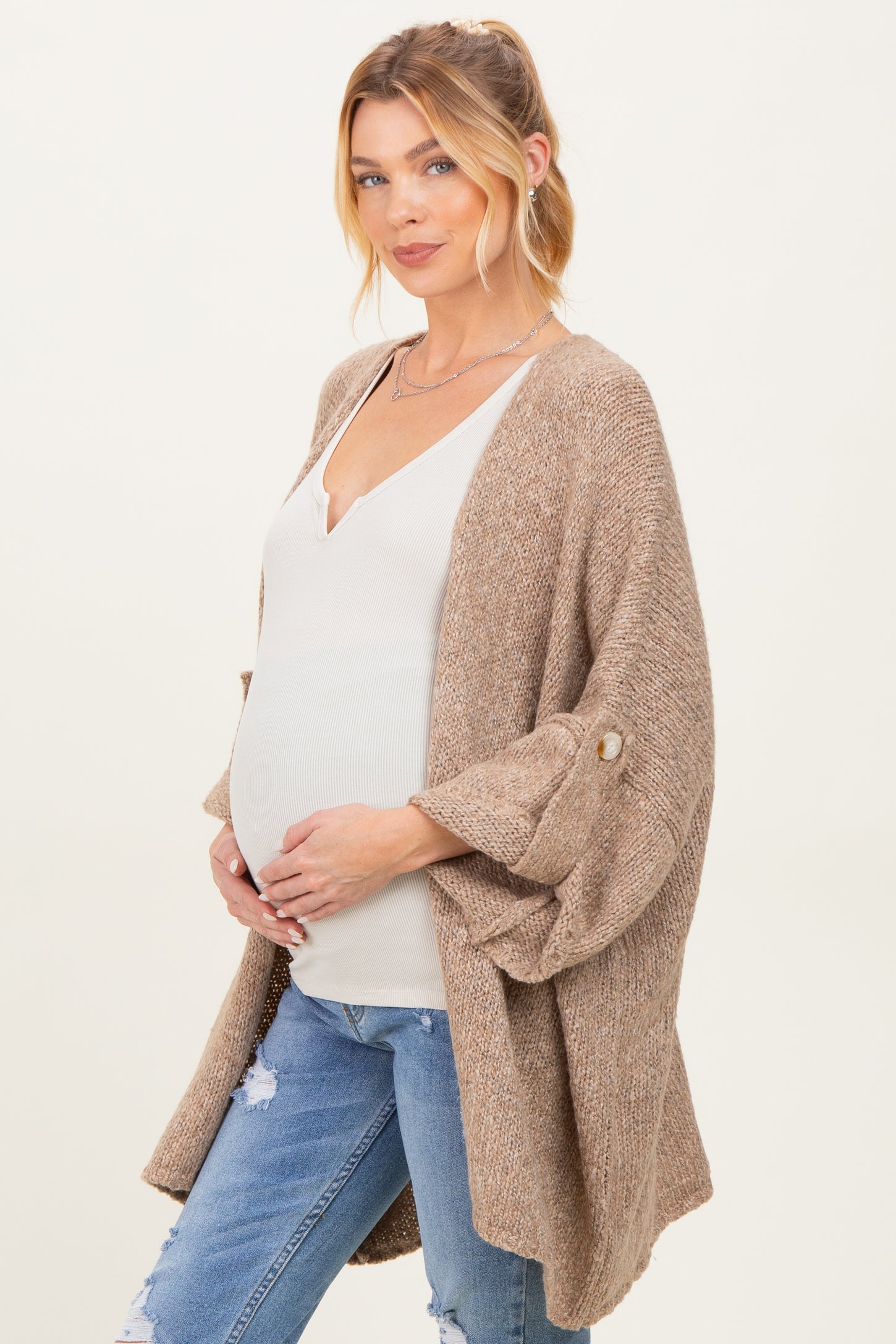 woman wearing a beige maternity cardigan over a white top and holding her baby bump with distressed jeans