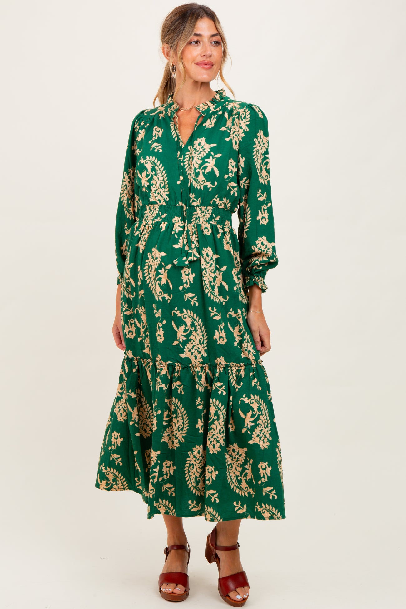 woman wearing a green floral maternity dress with long sleeves and a tiered skirt, smiling at the camera