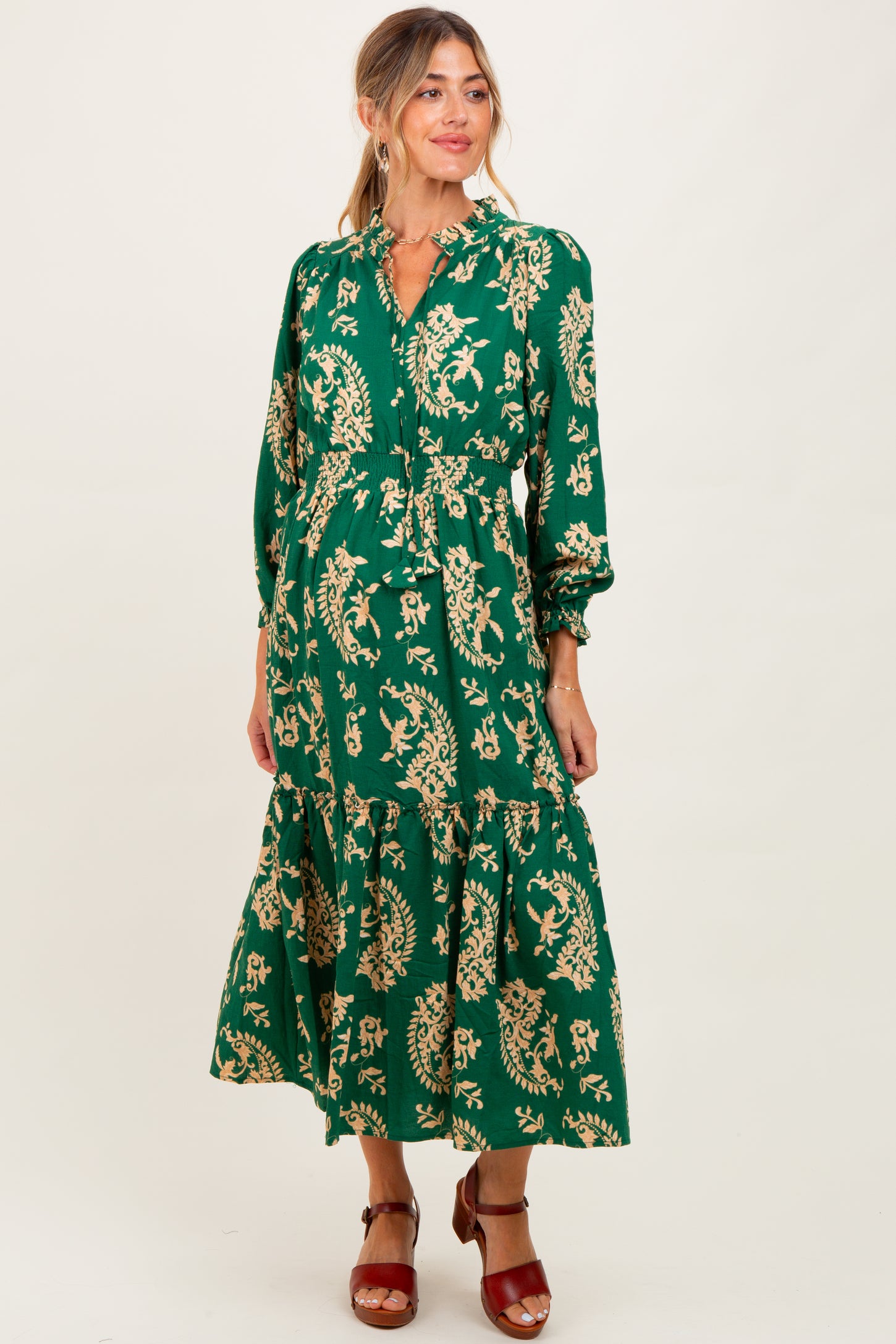woman wearing a green floral maternity dress with long sleeves and a tiered skirt, smiling at the camera