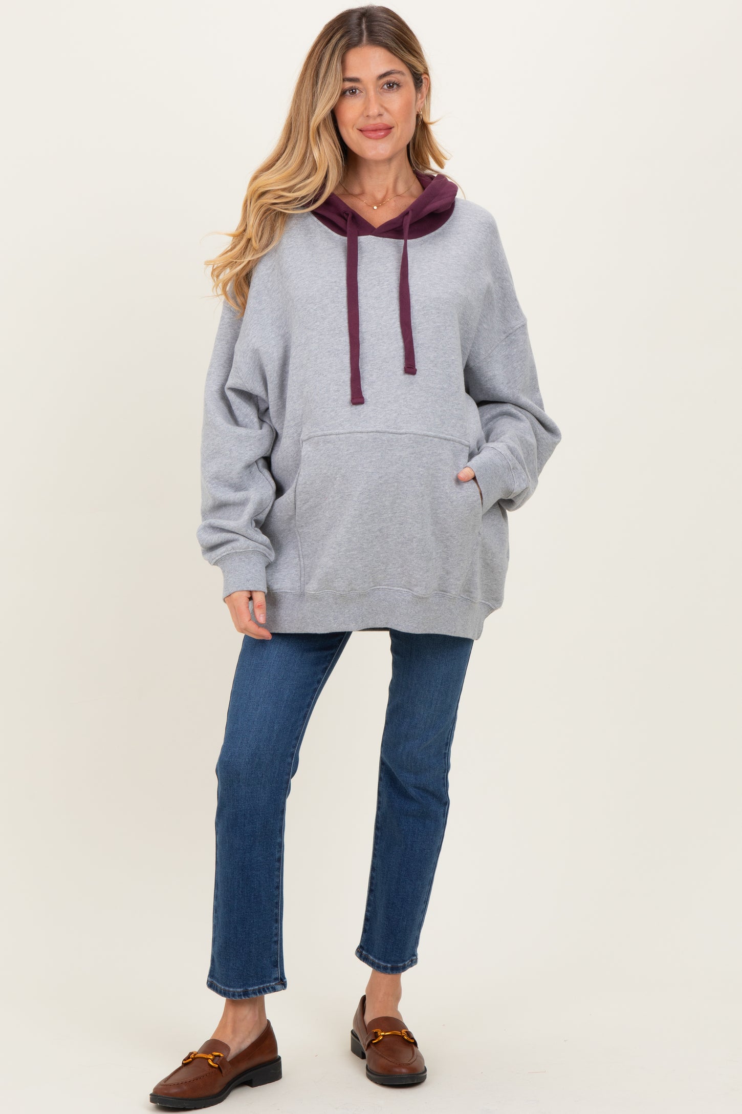 woman wearing a gray oversized maternity hoodie with burgundy accents and front pocket, paired with blue jeans