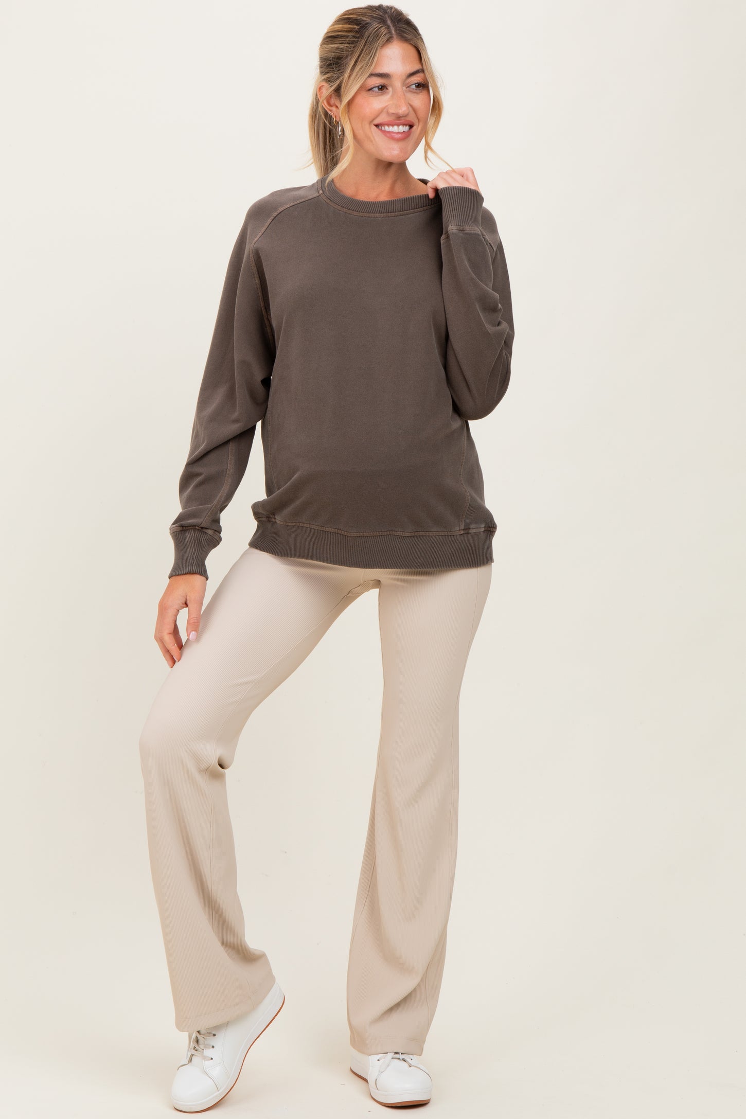 woman wearing a brown maternity sweatshirt and beige flared pants, smiling and posing with one hand on her hip
