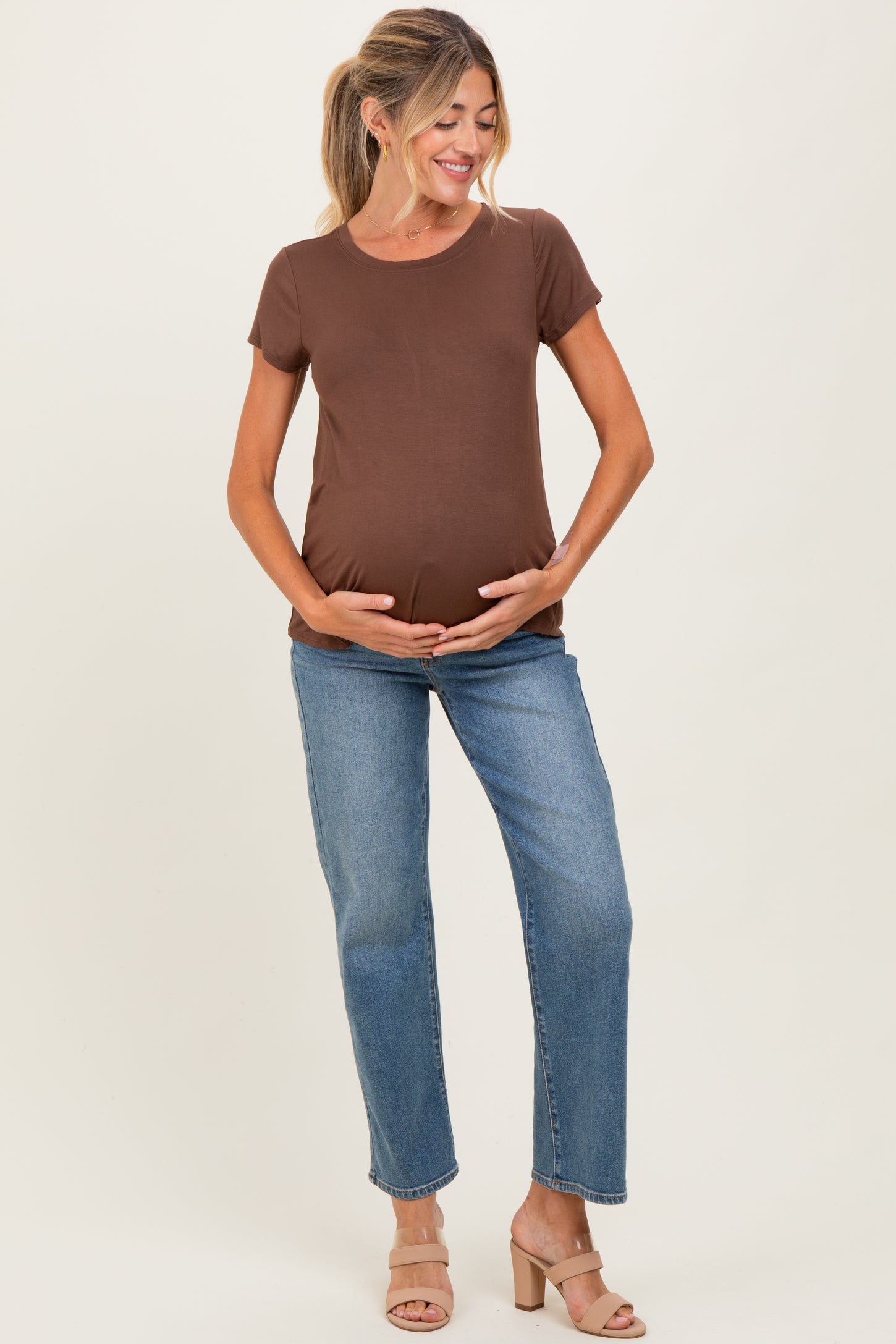 woman wearing a brown short sleeve maternity top and blue jeans, holding her baby bump