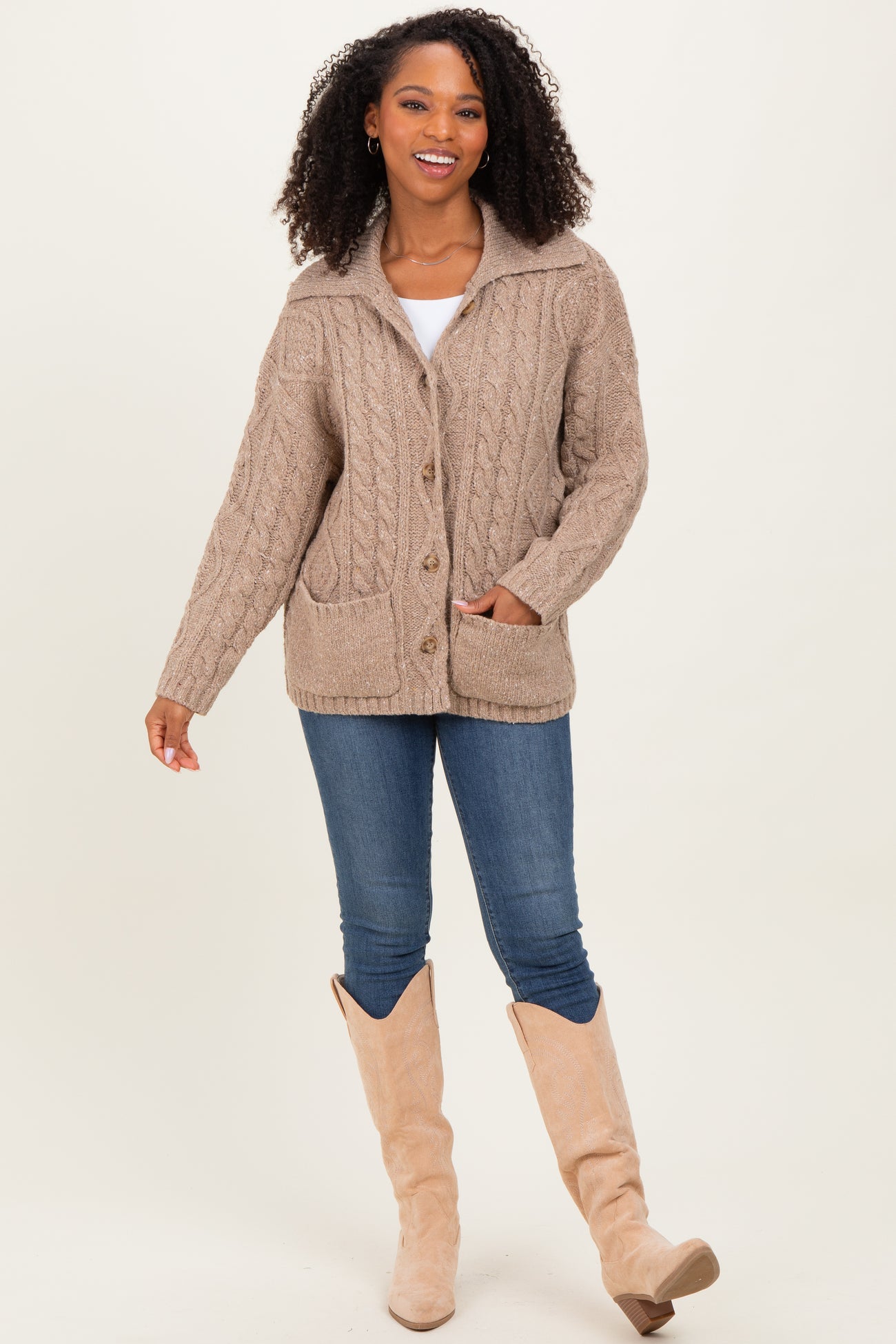 woman wearing a beige chunky cable knit cardigan with a collar, paired with blue jeans and beige over-the-knee boots