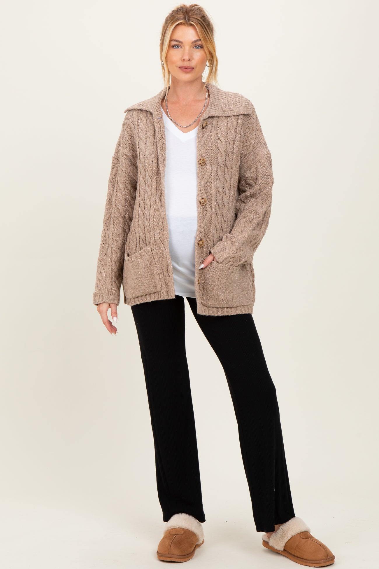 woman wearing a beige cable knit cardigan over a white top and black pants, displaying her baby bump