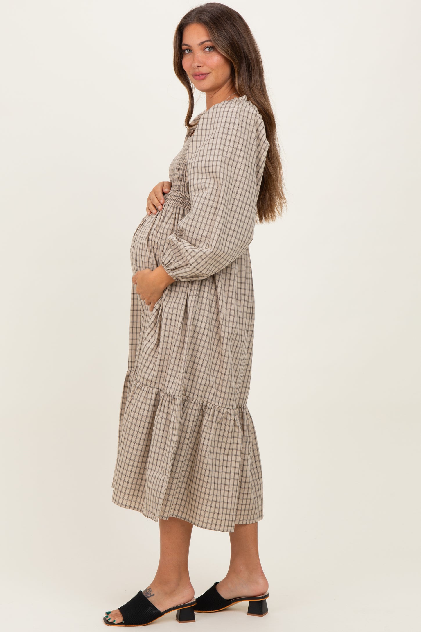 woman wearing a beige and white checkered maternity dress with long sleeves and ruffle hem, holding her baby bump