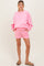 woman wearing a pink maternity sweatshirt and matching shorts, standing with hands in pockets