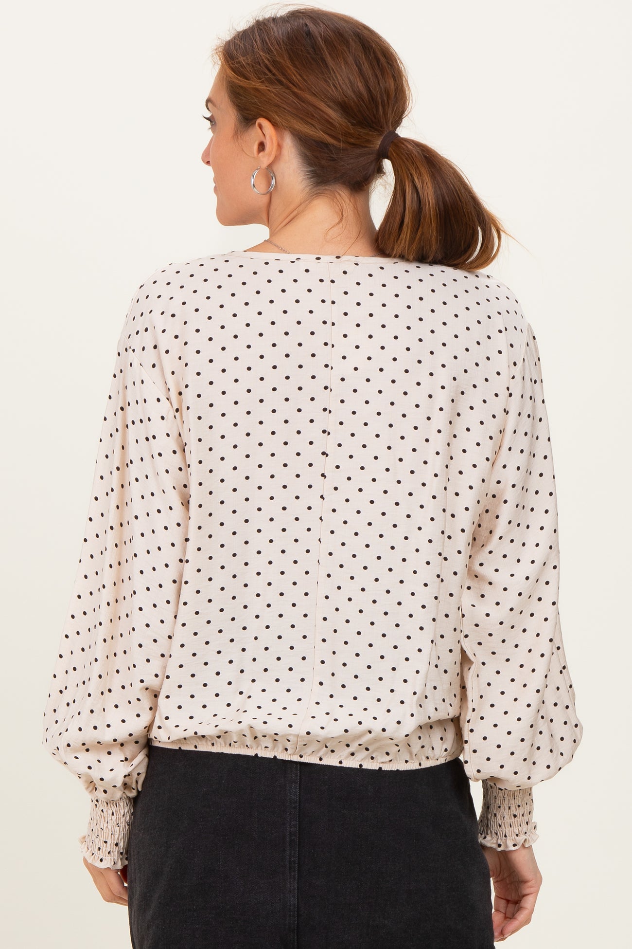 woman wearing a beige long sleeve top with black polka dots and a smocked hem, showing the back view