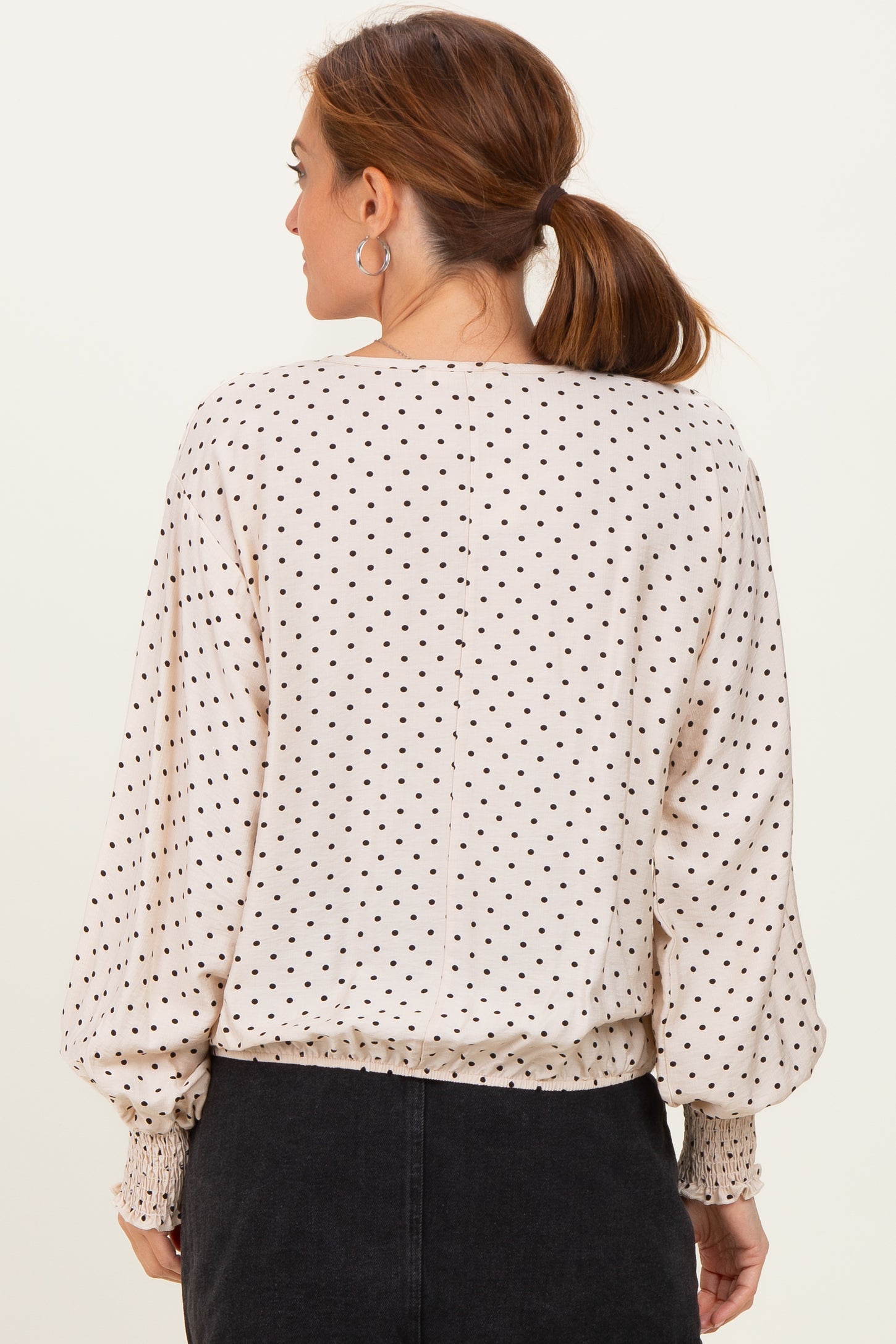 woman wearing a beige long sleeve top with black polka dots and a smocked hem, showing the back view