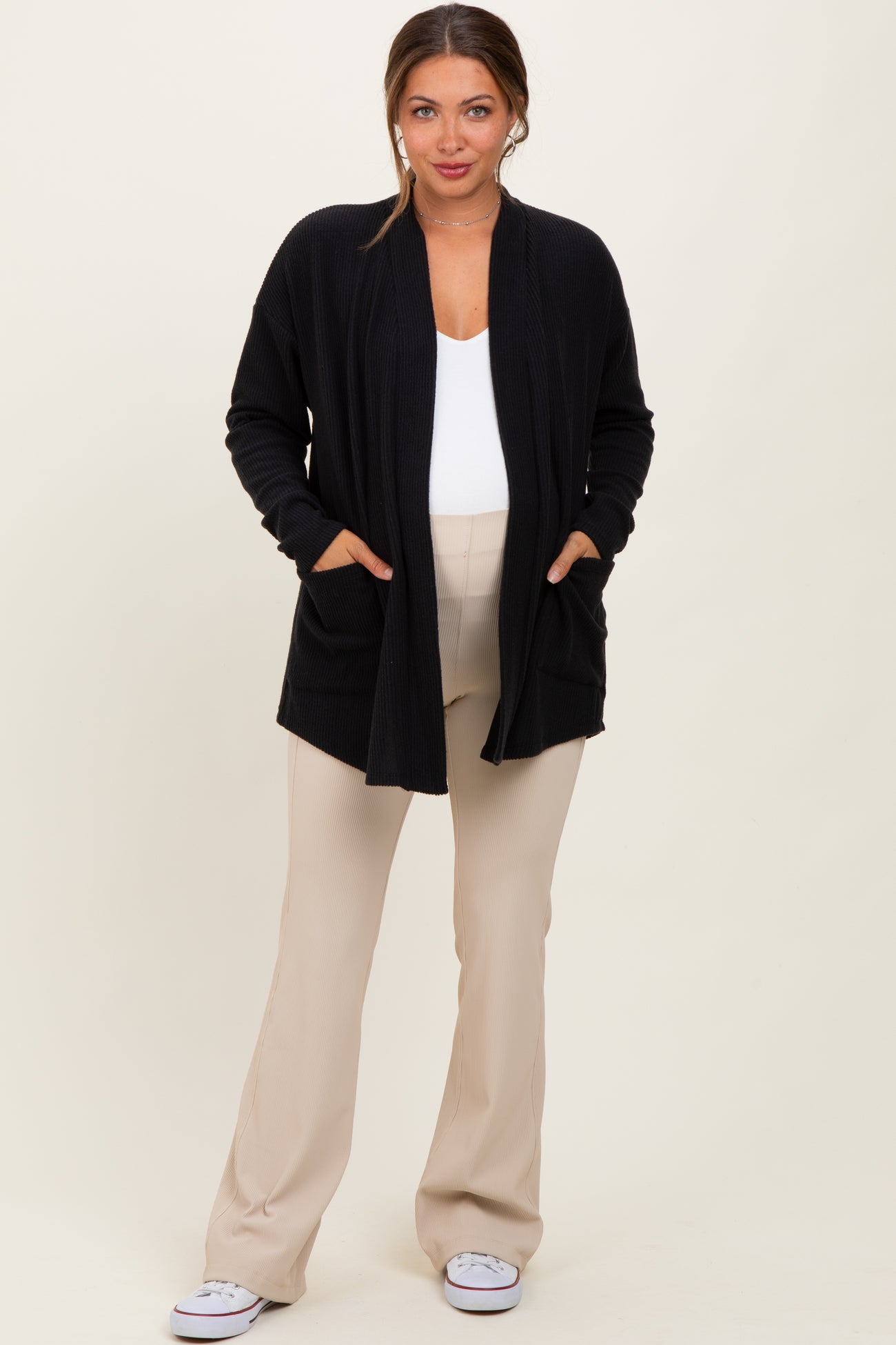woman wearing a black open front maternity cardigan with pockets over a white top and beige maternity pants, showing her baby bump