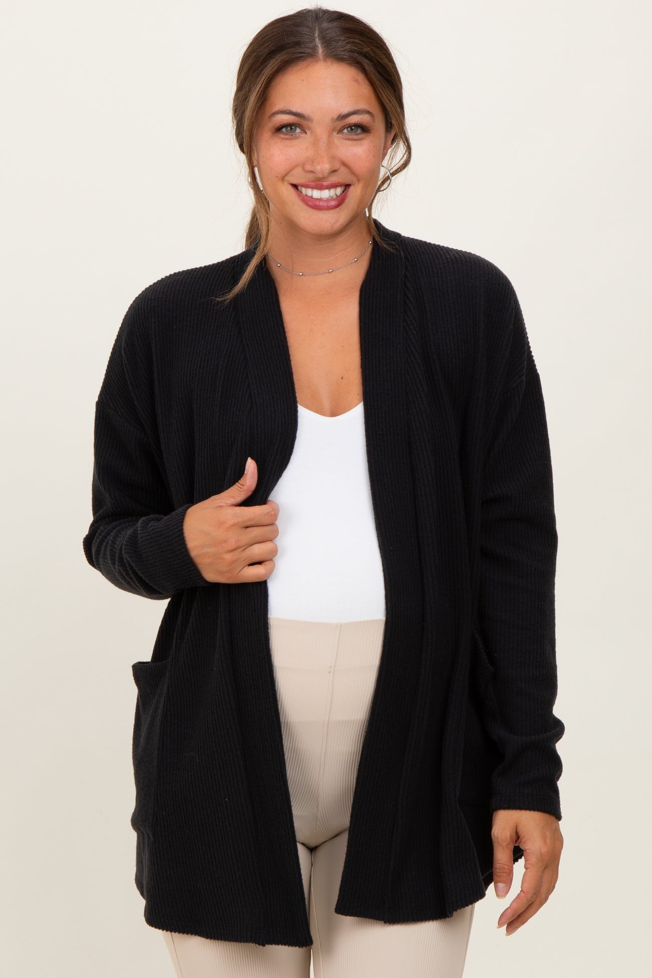woman wearing a black ribbed maternity cardigan over a white tank top and beige pants, smiling at the camera