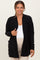 woman wearing a black ribbed maternity cardigan over a white tank top and beige pants, smiling at the camera