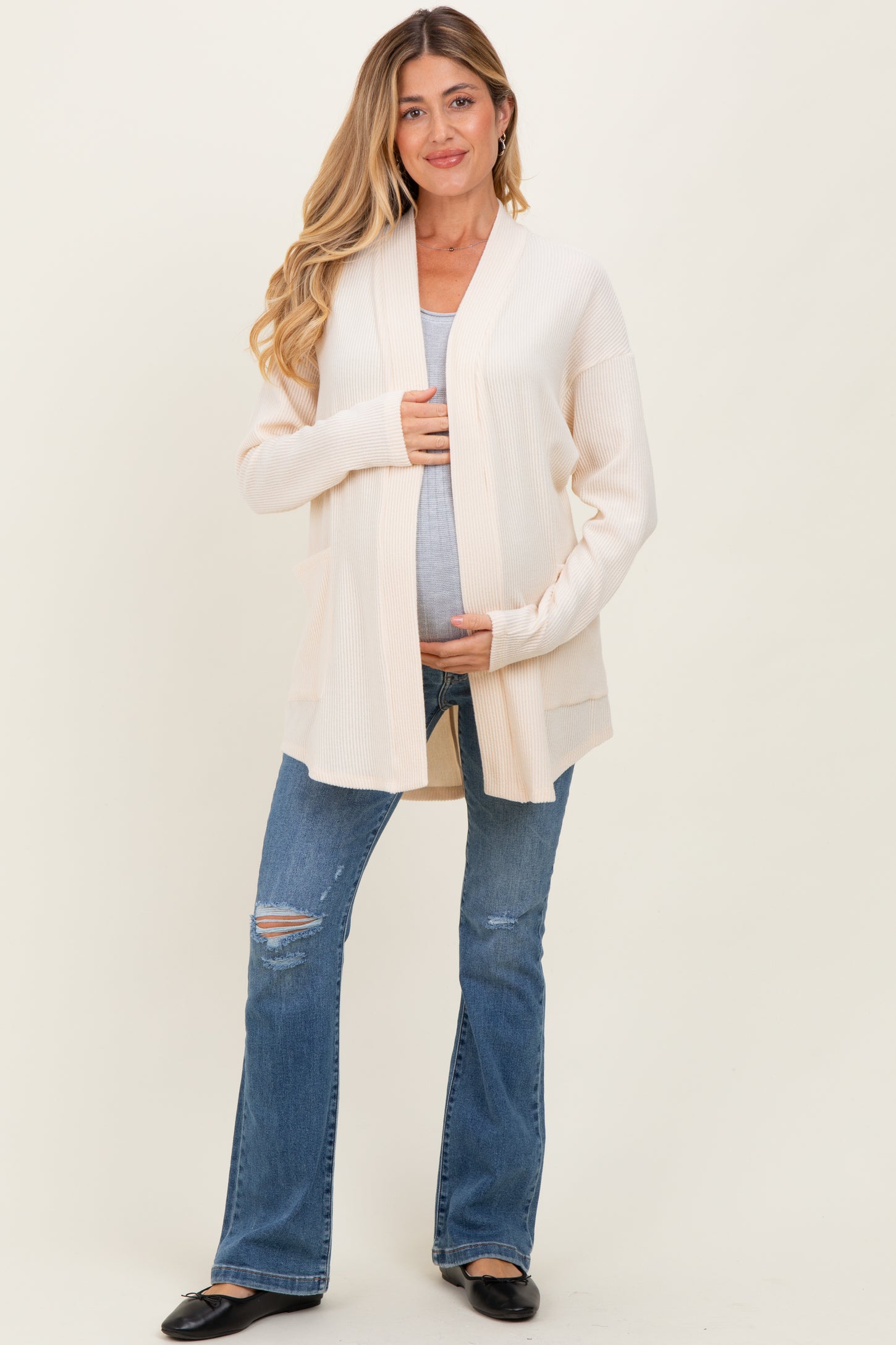 woman wearing a beige long cardigan over a tank top and holding her baby bump while standing in distressed blue jeans