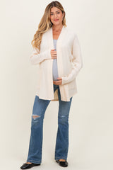 woman wearing a beige long cardigan over a tank top and holding her baby bump while standing in distressed blue jeans