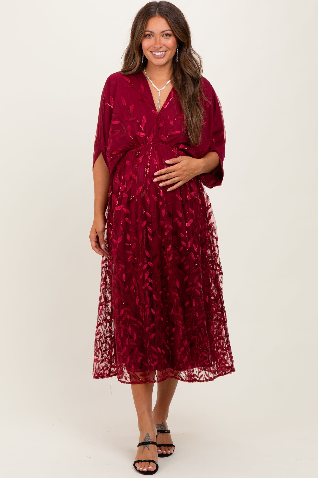woman wearing a burgundy maternity dress with floral embroidery and a flowy overlay, holding her baby bump