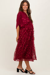 woman wearing a burgundy maternity dress with a floral lace overlay and a flowing silhouette, showcasing her baby bump