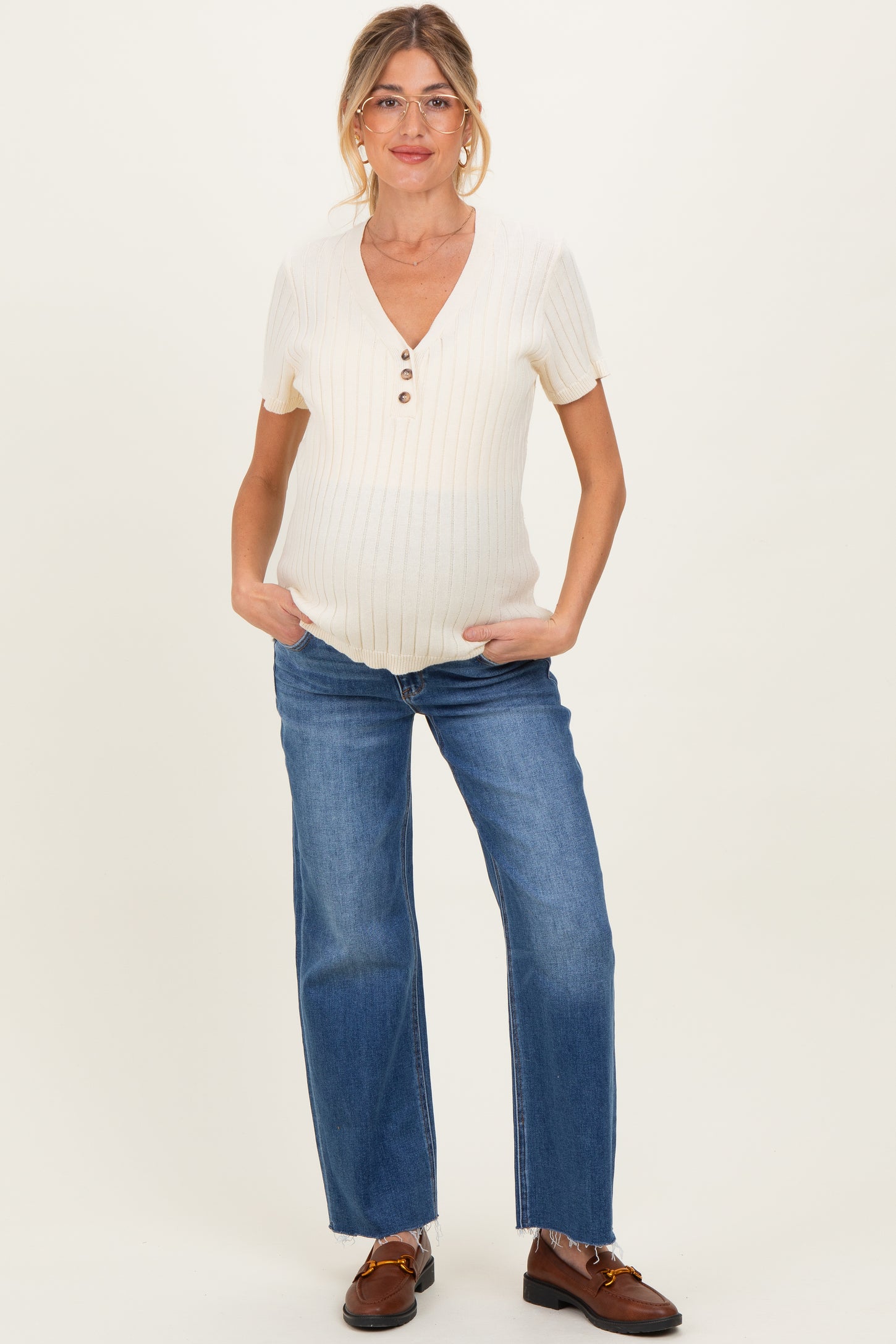 woman wearing a cream short sleeve maternity top with buttons and blue straight leg jeans, gently cradling her baby bump