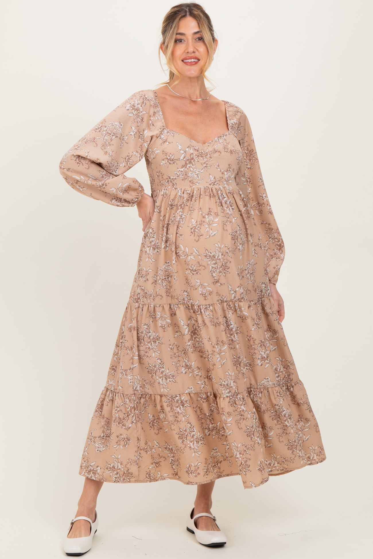 woman wearing a light brown floral maternity dress with long sleeves and a tiered skirt, highlighting her baby bump