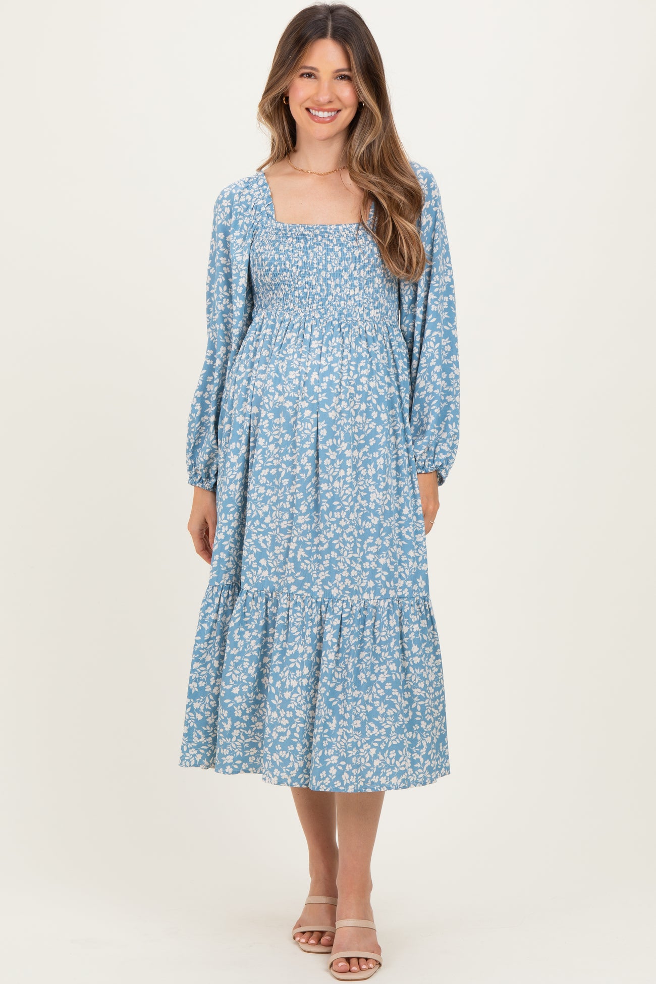 woman wearing a light blue floral maternity dress with a square neckline and long sleeves, posing with a visible baby bump