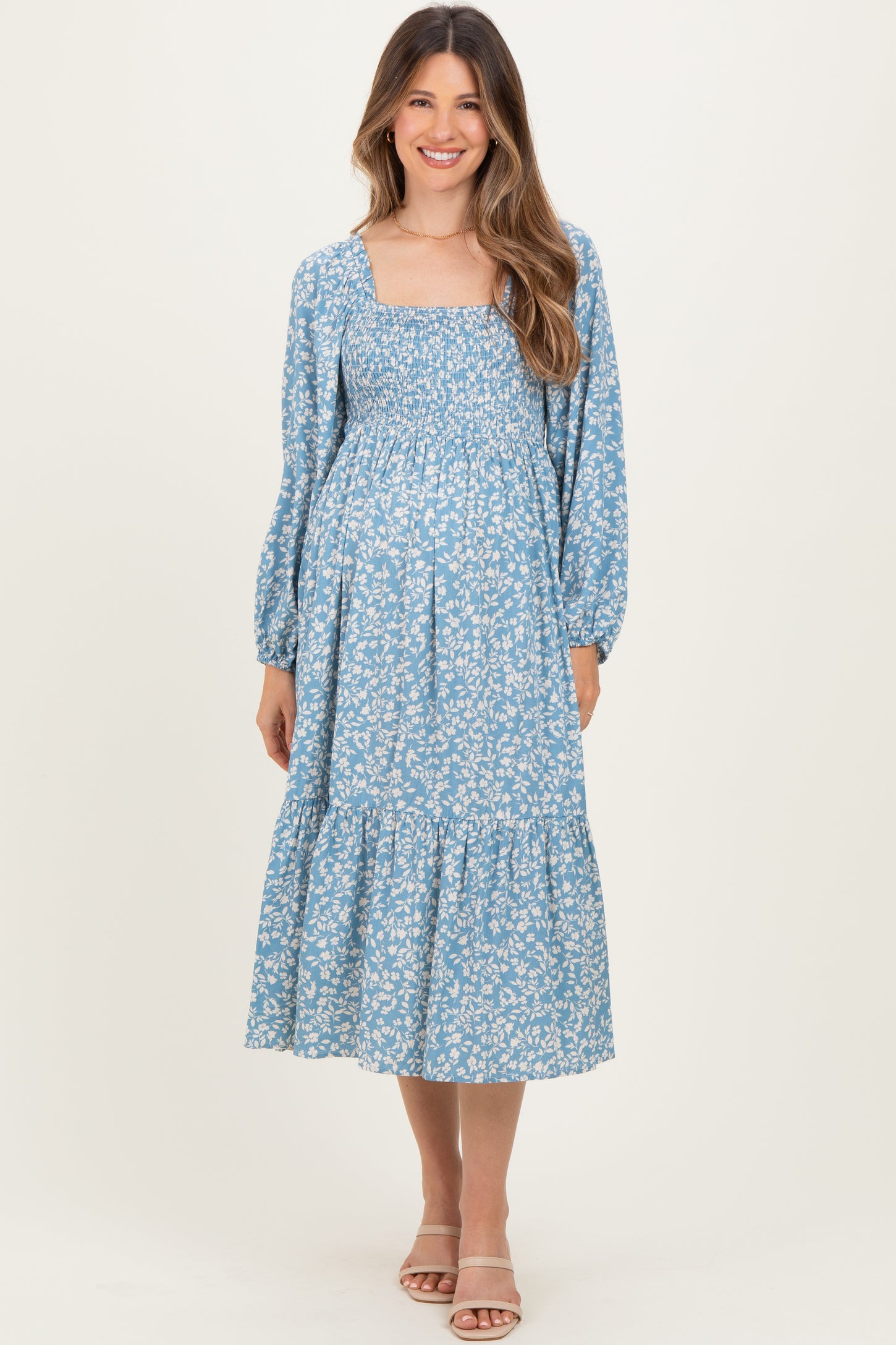 woman wearing a light blue floral maternity dress with a square neckline and long sleeves, posing with a visible baby bump