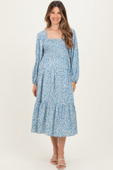 woman wearing a light blue floral maternity dress with a square neckline and long sleeves, posing with a visible baby bump
