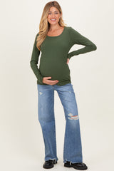 woman wearing an olive green long sleeve maternity top and distressed blue flared jeans, cradling her baby bump