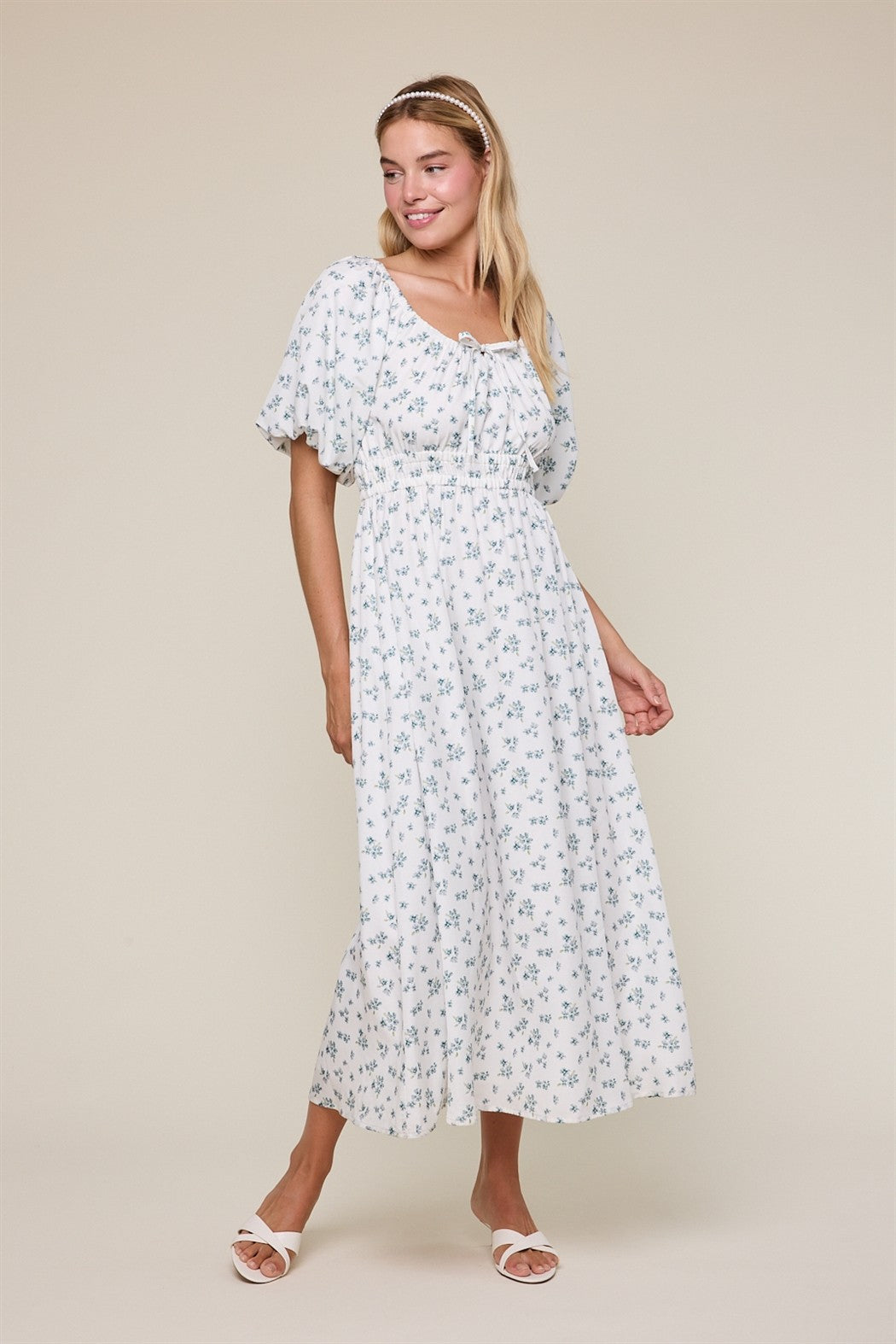 woman wearing a white floral maxi dress with short puff sleeves and a tie neckline, standing on a neutral background