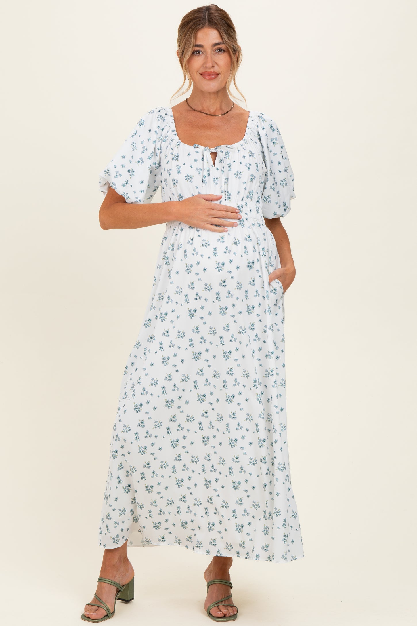 woman wearing a white floral maternity dress with puff sleeves and a baby bump, standing with one hand on her belly