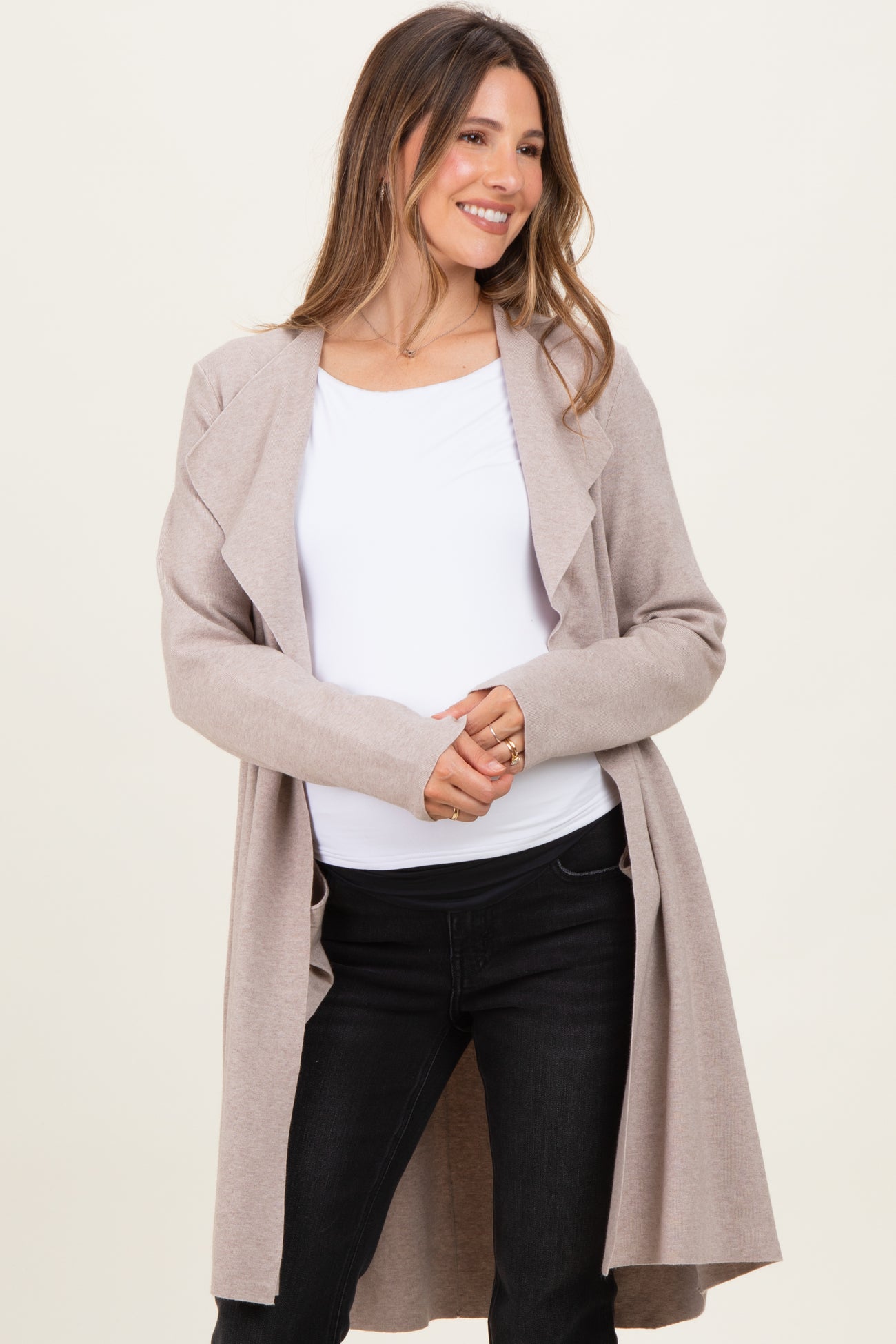 woman wearing a light beige maternity cardigan over a white top and black jeans, smiling with her hands on her waist