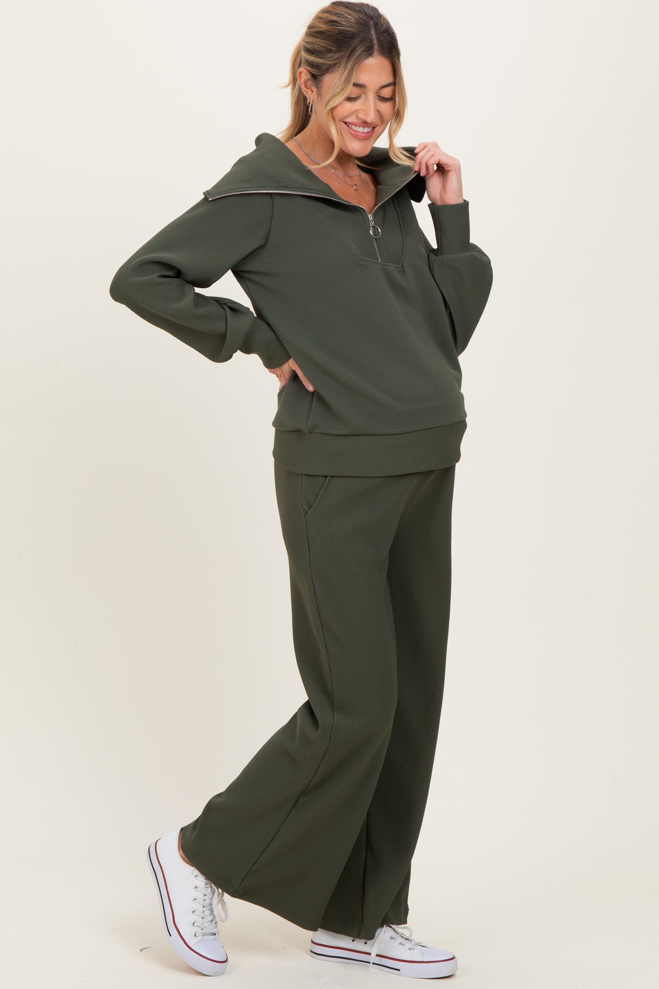 woman wearing a dark green maternity zip-up sweatshirt and matching wide-leg pants, styled with white sneakers