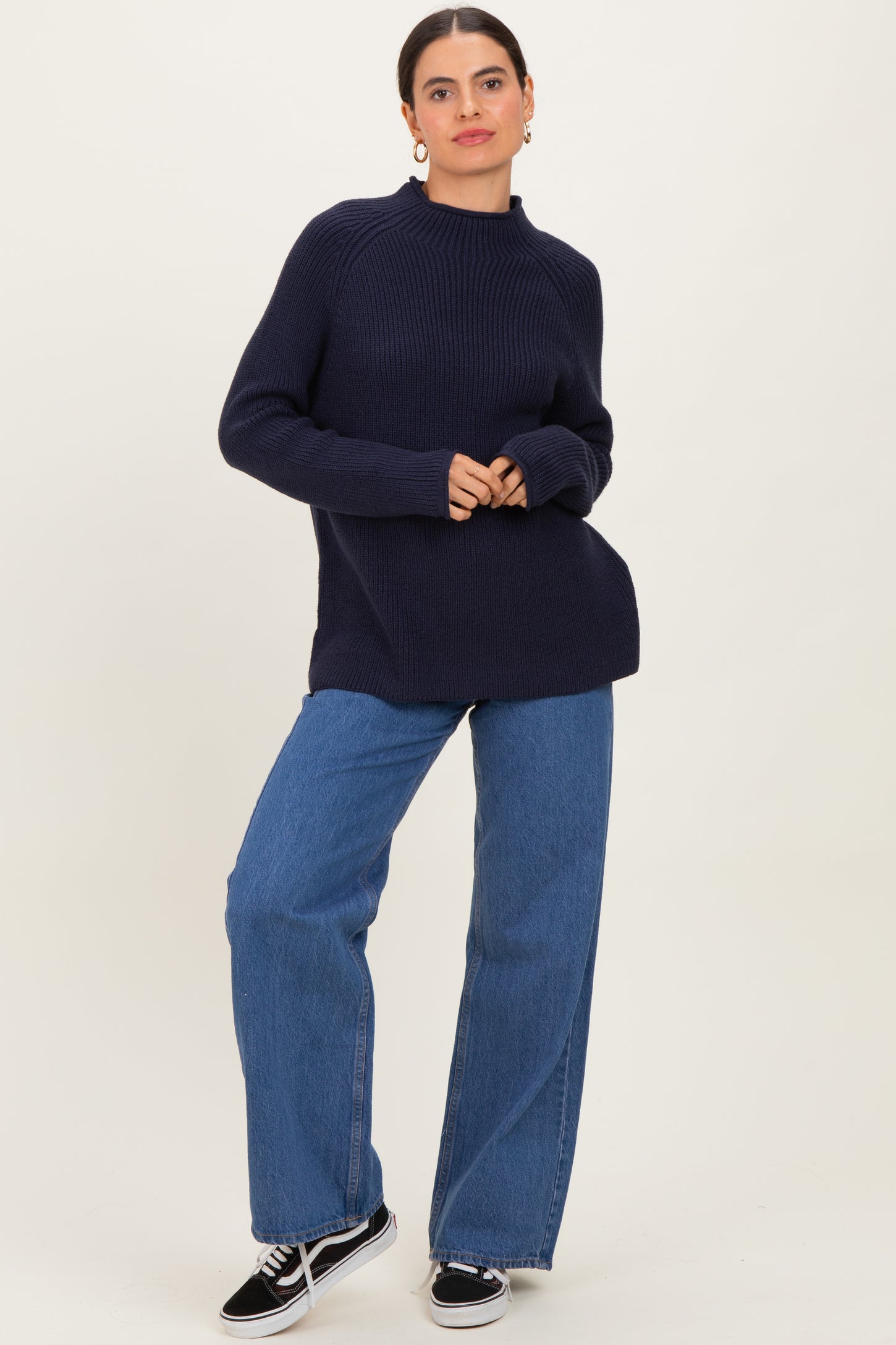 woman wearing a dark navy ribbed sweater and wide-leg blue jeans, standing with hands clasped in front