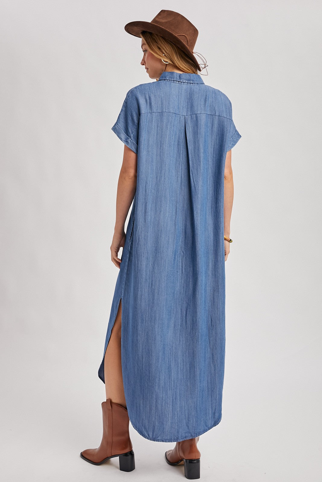 woman wearing a long denim shirt dress with short sleeves, side slits, and a collar, accessorized with a brown hat and boots
