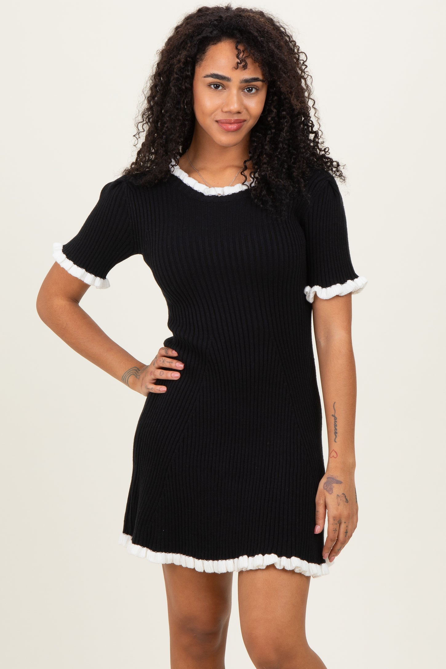 woman wearing a black ribbed dress with white ruffled sleeves and hem, posing with one hand on her hip
