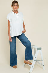 woman wearing a white maternity polo top with short sleeves and blue wide-leg jeans, standing next to a ladder