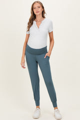 woman wearing a grey henley maternity top and teal jogger pants with a wide waistband and side pockets, showing her baby bump