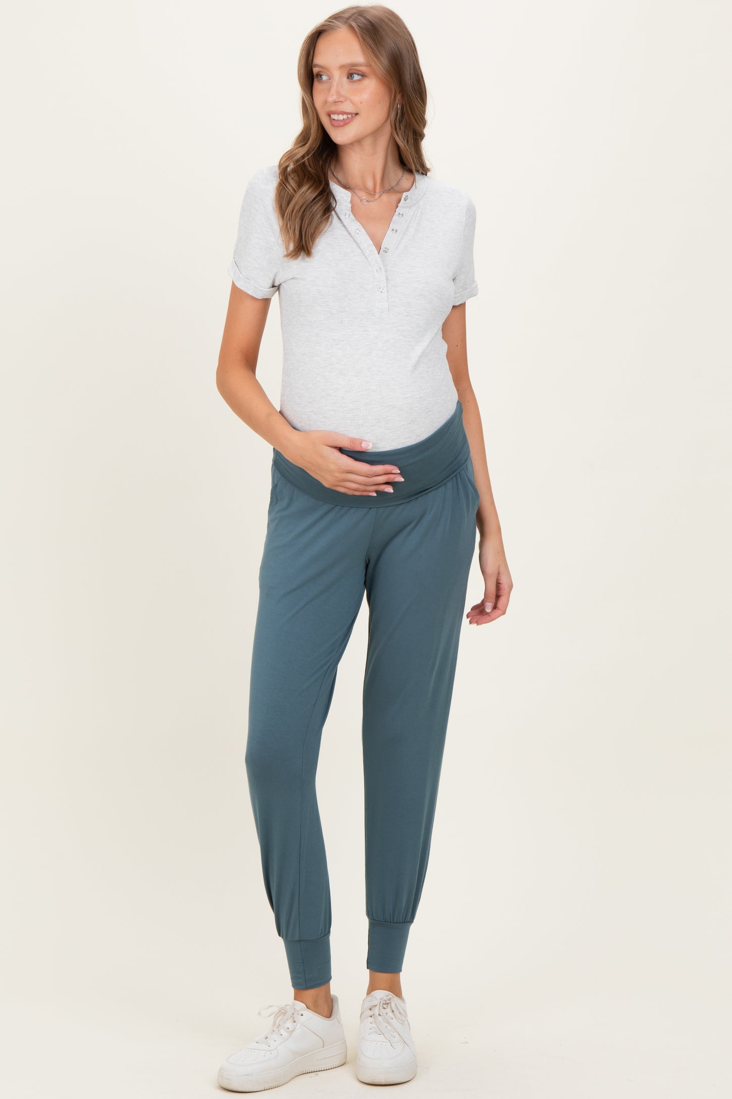 woman wearing a light gray maternity polo shirt and teal maternity joggers, holding her baby bump