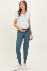 woman wearing a light gray maternity polo shirt and teal maternity joggers, holding her baby bump