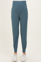 woman wearing teal maternity jogger pants with a high waistband and pockets, paired with white sneakers