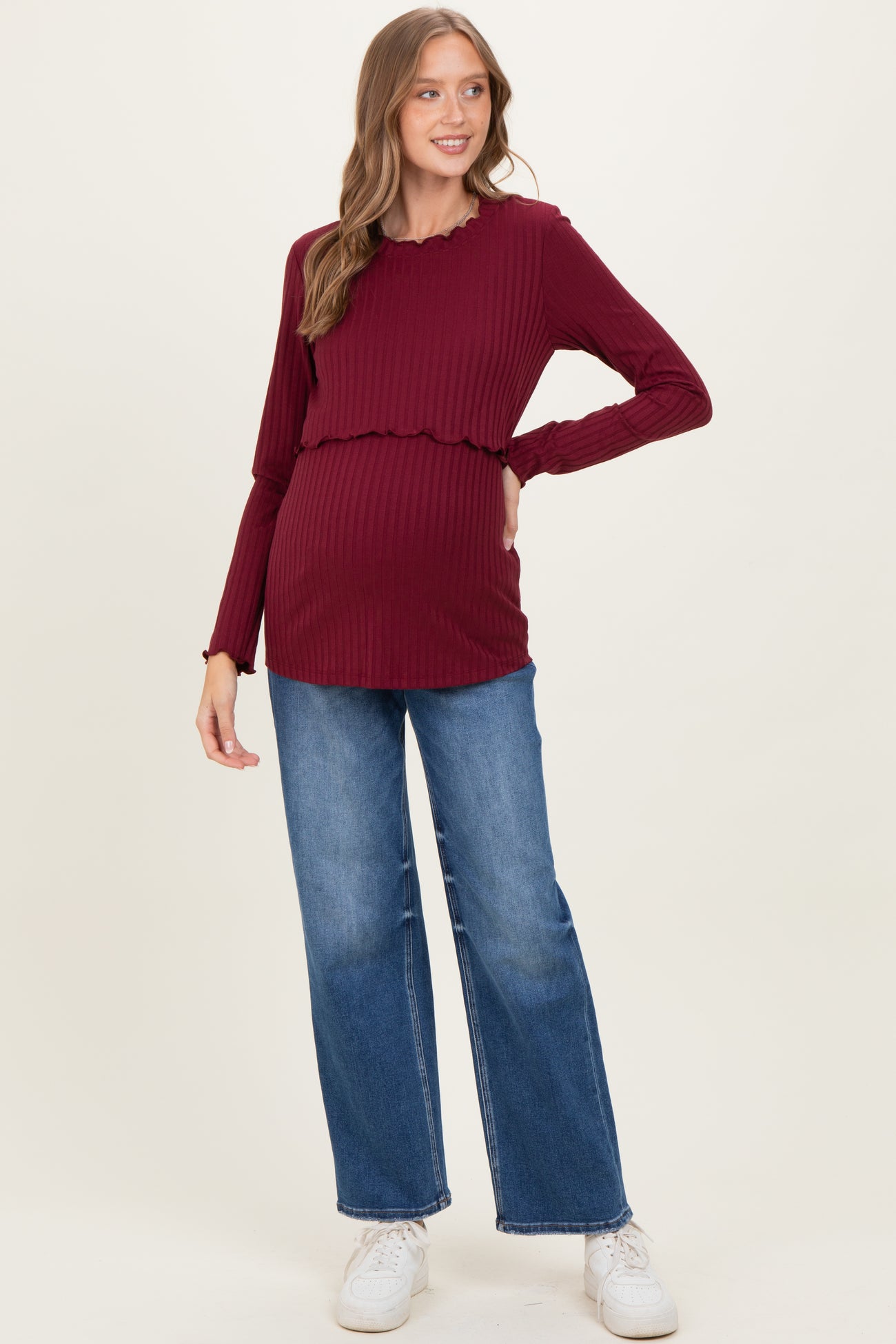 woman wearing a burgundy ribbed maternity top with a scalloped hem and a visible baby bump paired with blue wide leg jeans