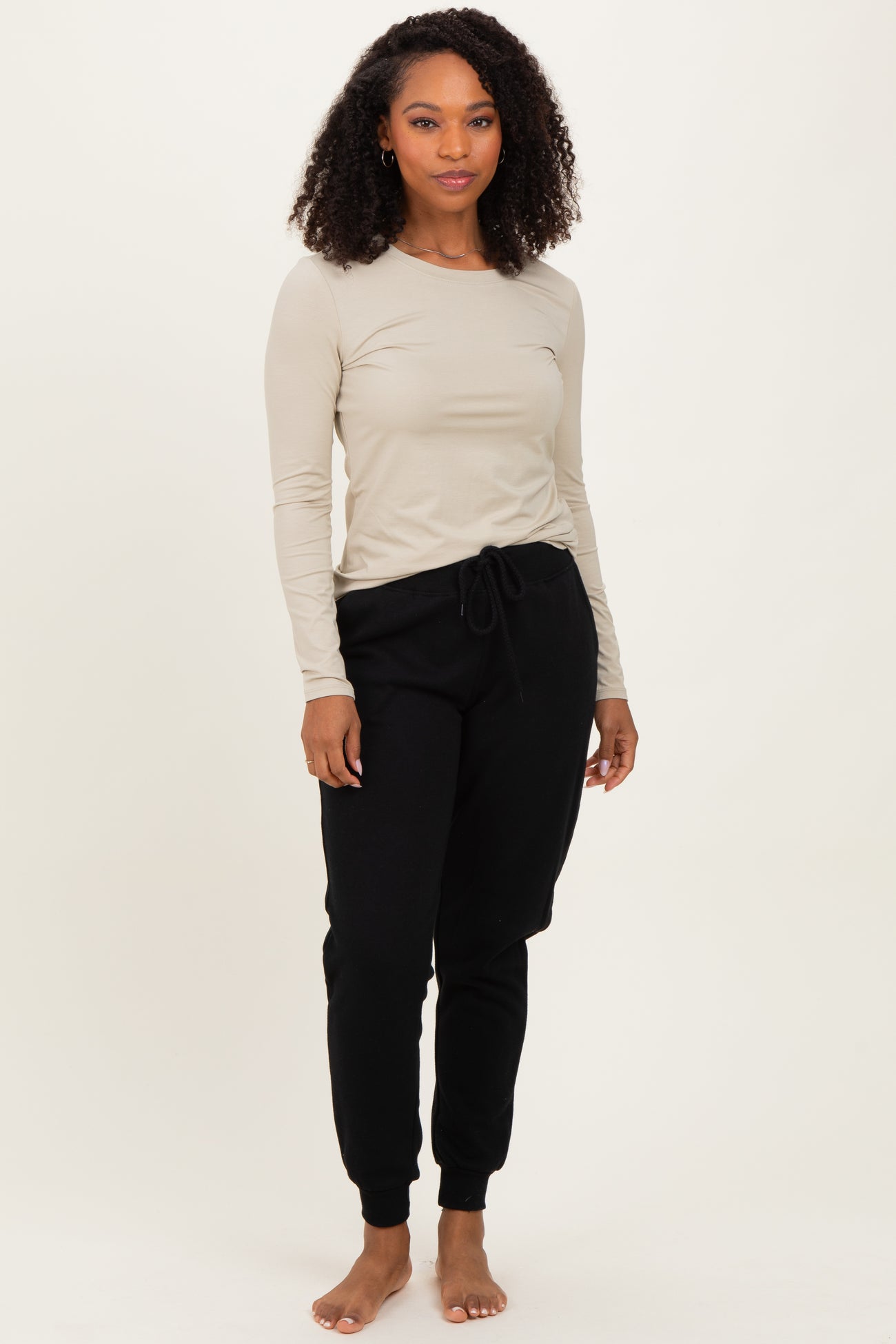 woman wearing a beige long sleeve top and black drawstring sweatpants, standing barefoot against a light background