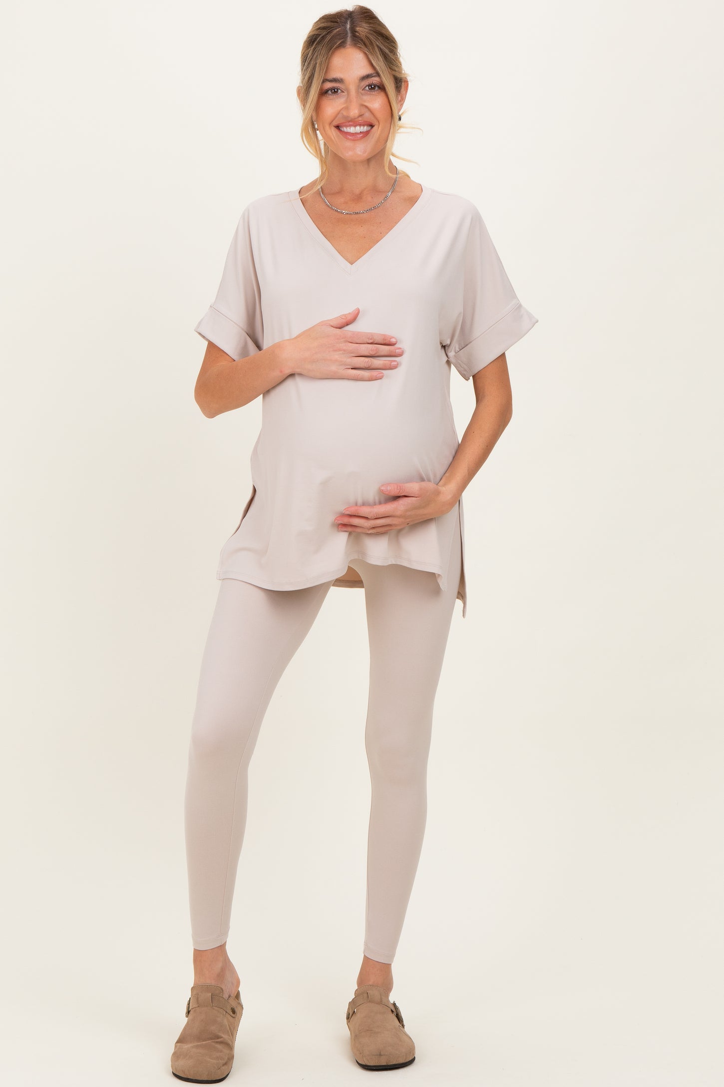 woman wearing a beige maternity top and matching leggings, holding her baby bump with a smile