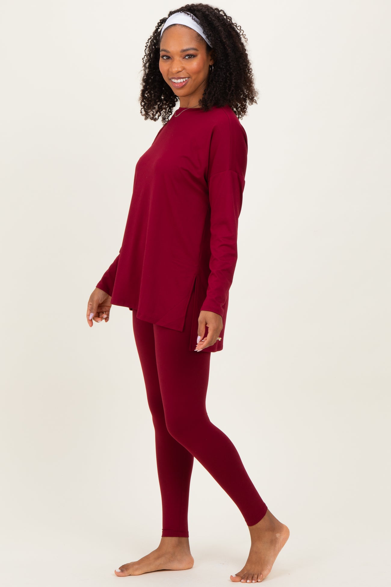 woman wearing a burgundy long sleeve top and matching leggings, standing barefoot with a slight smile