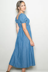 woman wearing a blue denim maxi dress with puff sleeves and a gathered back detail, smiling over her shoulder