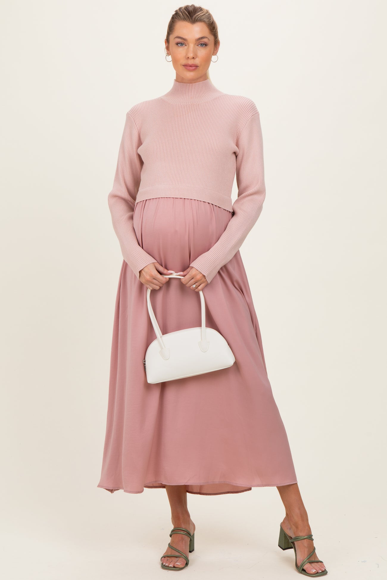 woman wearing a light pink ribbed maternity sweater and a flowy mauve maxi skirt, holding a white handbag and showing her baby bump
