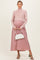 woman wearing a light pink ribbed maternity sweater and a flowy mauve maxi skirt, holding a white handbag and showing her baby bump