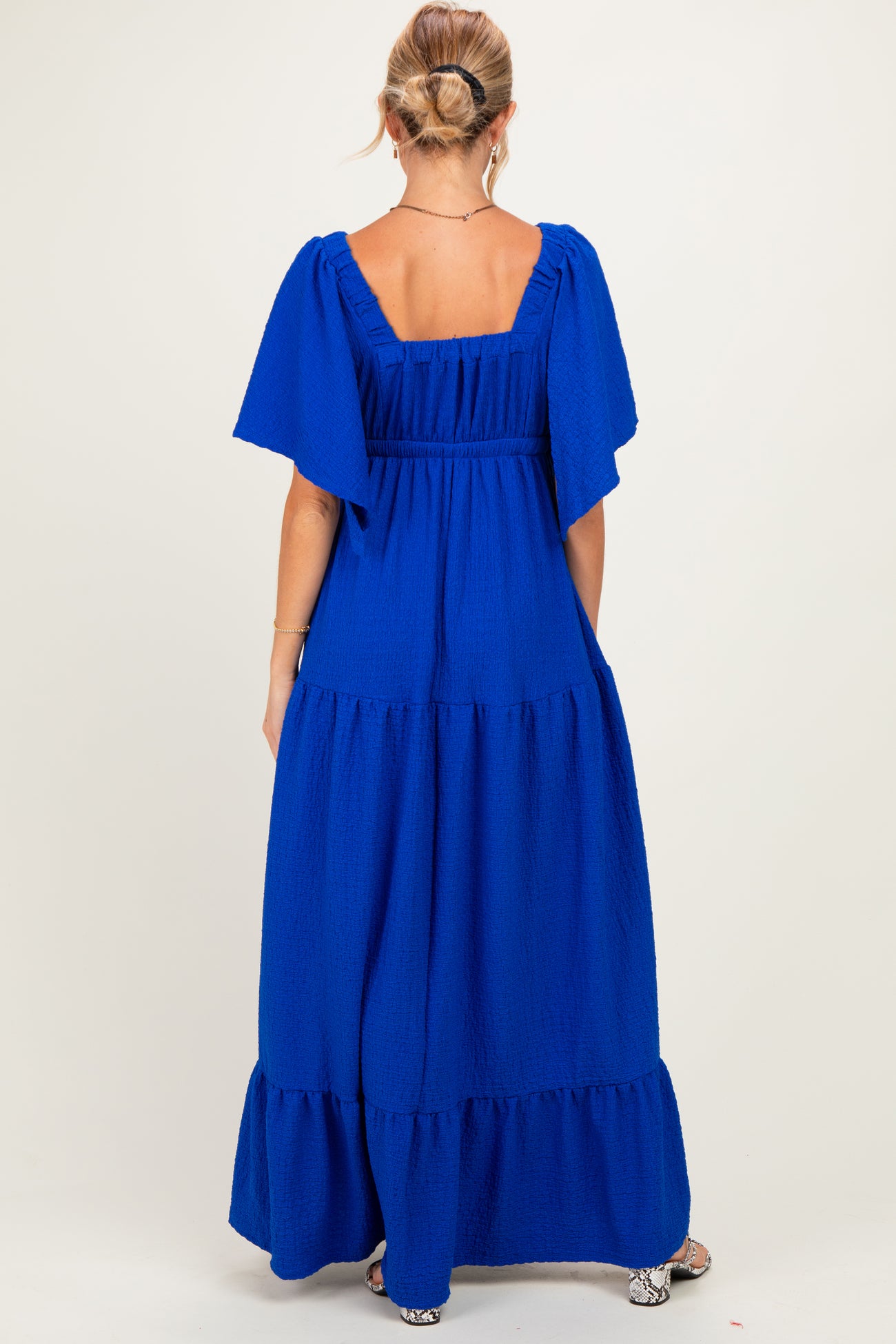 woman wearing a bright blue maternity maxi dress with flutter sleeves and a tiered skirt, showing the back view